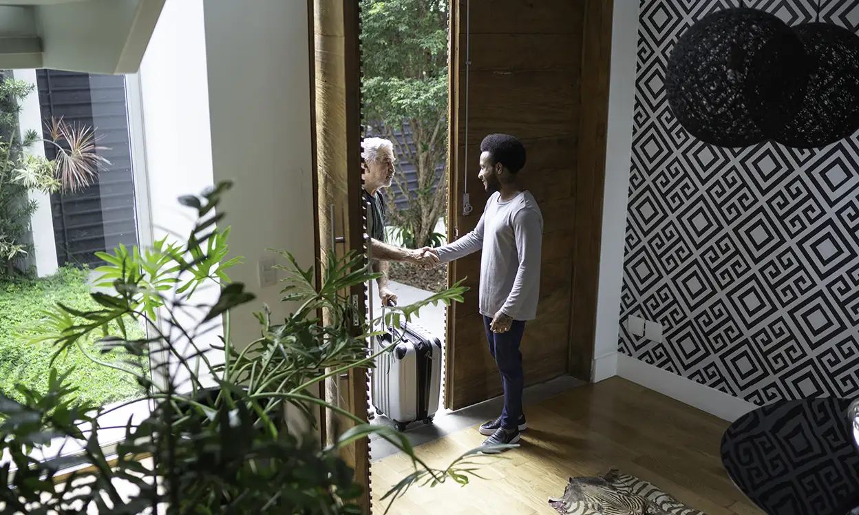 man welcoming man with suitcase into his house